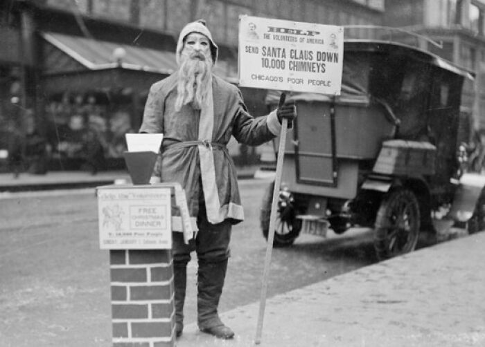 Creepy Santa holding a sign about sending Santa down chimneys, standing next to a collection box on a city street.