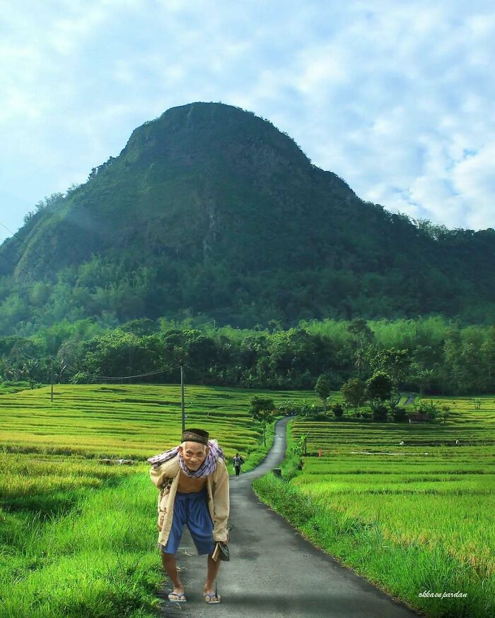 Indonesian photographer captures a local carrying a sack on a rural path with lush green fields and a mountain backdrop.