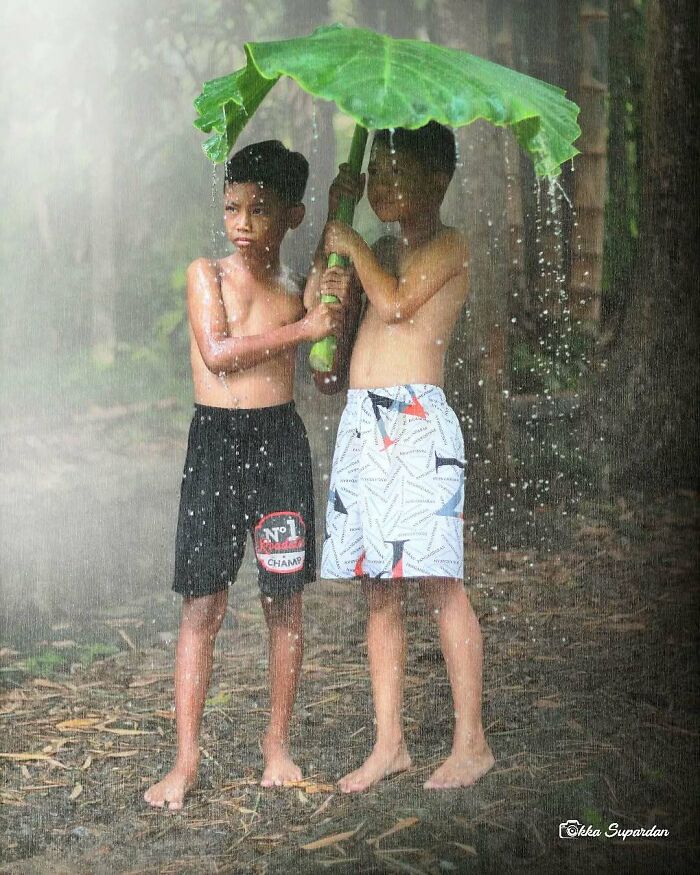 Two boys in rain using a large leaf as an umbrella, captured by an Indonesian photographer highlighting simple life moments.