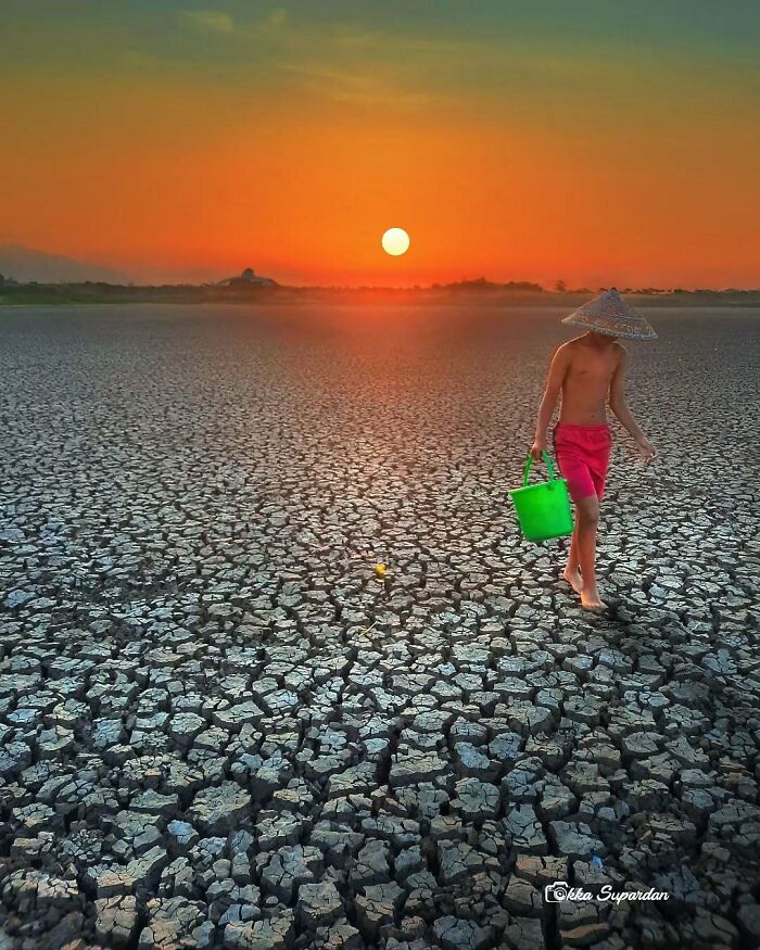 Indonesian photographer captures a child walking on c*****d earth at sunset, holding a green bucket, depicting simple life moments.
