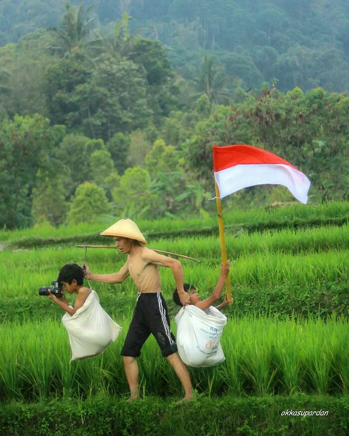 Indonesian photographer captures simple life moments; a man carries children in bags through lush fields, holding a flag.