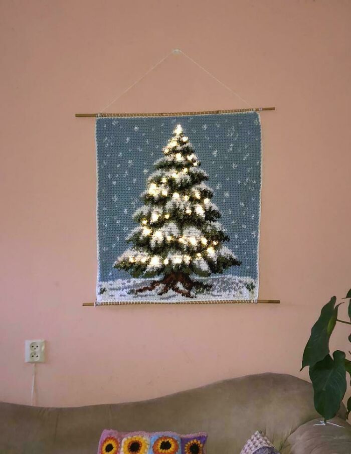 Crochet enthusiasts showcase a wall hanging of a lighted Christmas tree with snow on a blue background.