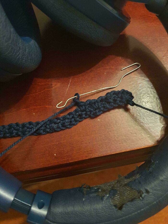 Close-up of a small navy blue crochet project and a wire crochet hook resting on a wooden surface near worn blue headphones.