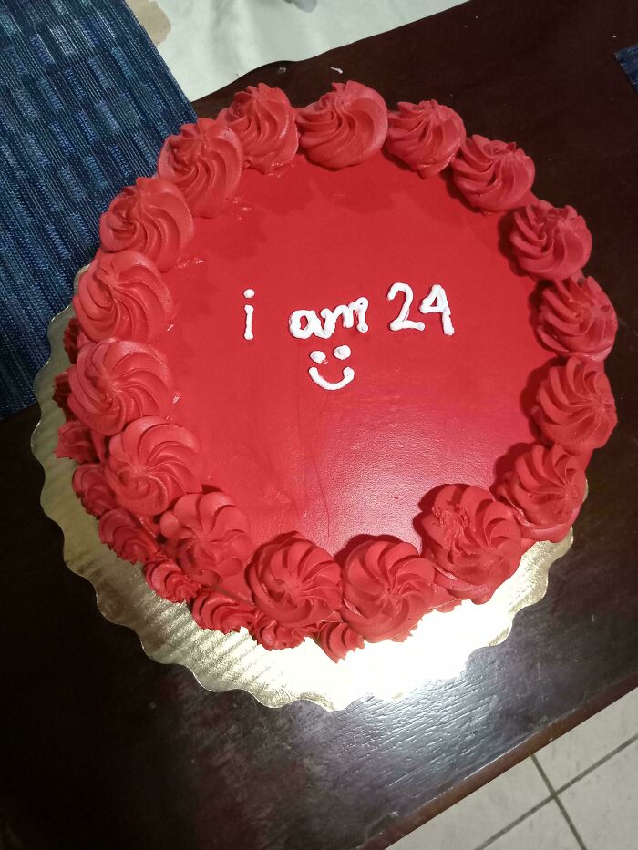 Red cake with frosting flowers, text "I am 24" in white, and smiley face. Uninteresting decoration.