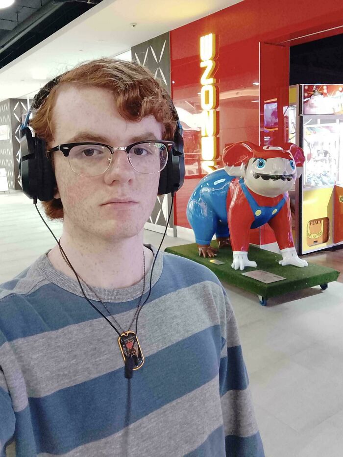 Person with headphones poses in front of a colorful whimsical statue at a mall; nothing interesting happening.