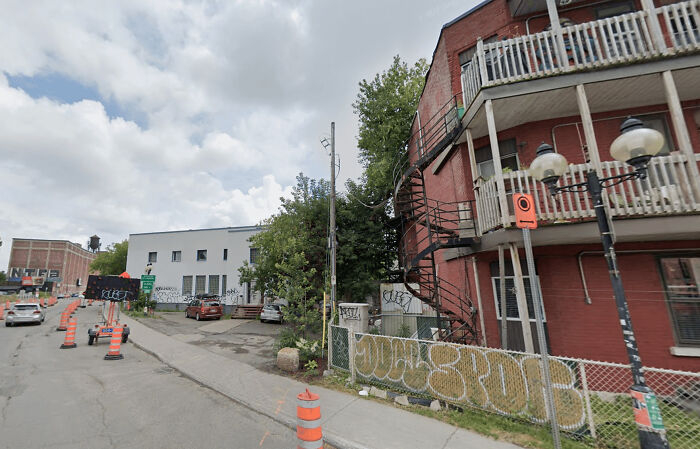 Urban hell scene with construction cones, graffiti, and a red brick building featuring spiral stairs and cluttered balconies.
