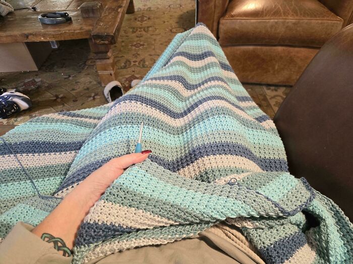 Hand holding crochet hook working on a striped blue and white crochet blanket in a cozy living room.