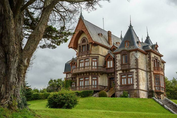 Victorian stone mansion with intricate wooden balconies and steep slate roof surrounded by lush greenery, showcasing impressive architectural feats.
