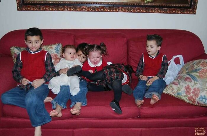 Awkward family photo of five children sitting closely on a red couch, with the middle child visibly upset.