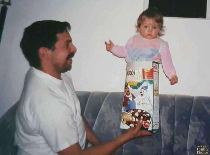 Awkward family photo of a man holding a baby in a cereal box on a sofa.