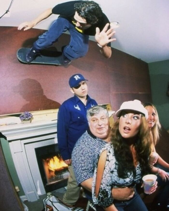 Skateboarder defying gravity indoors, over a surprised group by a fireplace, showcasing weirdness.