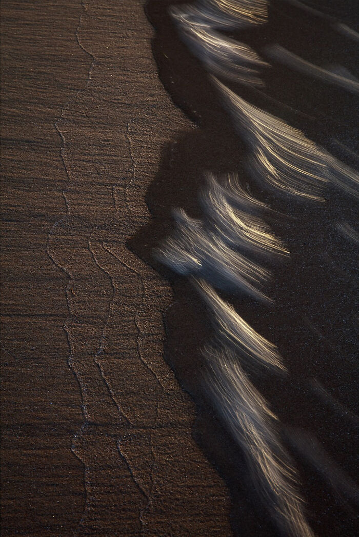 Photographer captures mesmerizing waves on a dark sandy beach, highlighting their delicate patterns and movement.