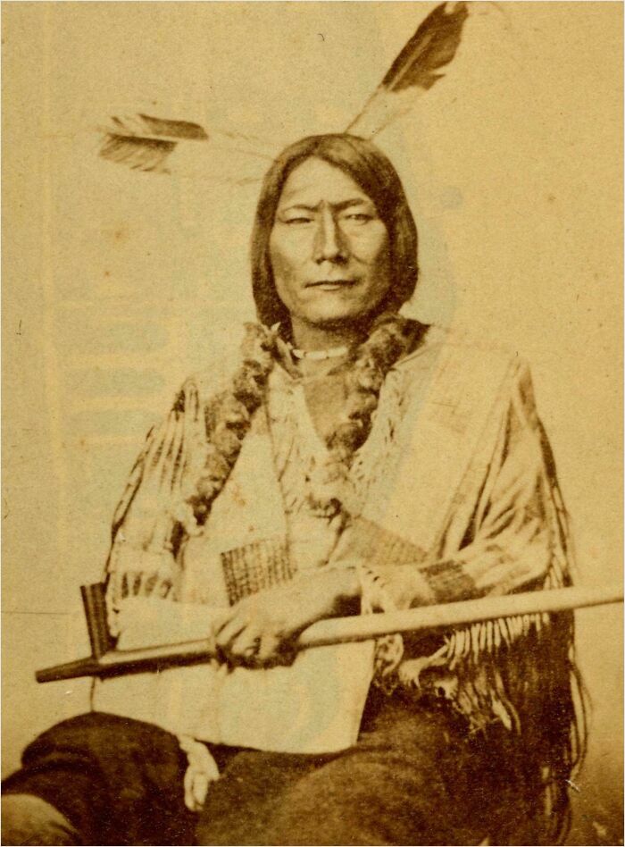A historical photo of a Native American man with braided hair wearing traditional attire and holding a staff.