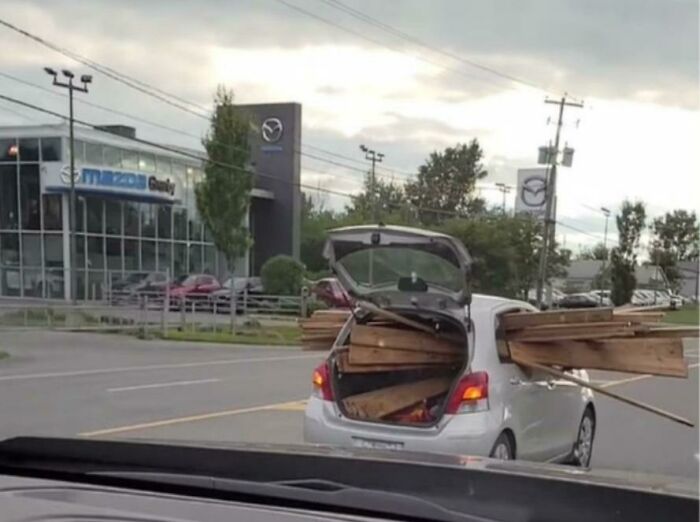 Car with open trunk overloaded with wooden planks on a street near a car dealership.