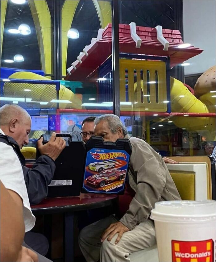 Three men sitting in a McDonald's play area, looking at a Hot Wheels case, resembling a potential album cover concept.