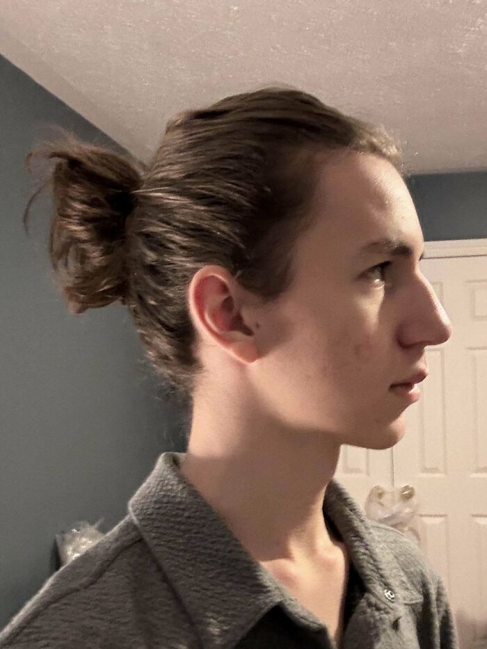 Young man with fierceflow beautiful hair tied in a messy bun, wearing a gray shirt indoors with a blue wall background.