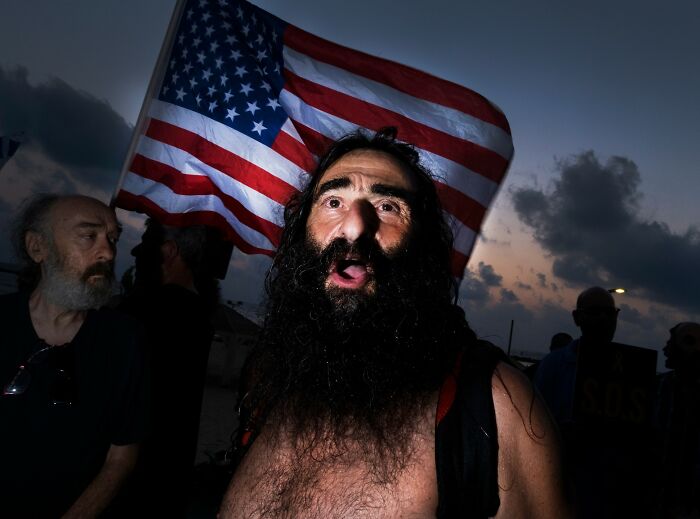 Man with a beard stands in front of an American flag at sunset, captured in Ilan Ben Yehuda's street photography in Israel.