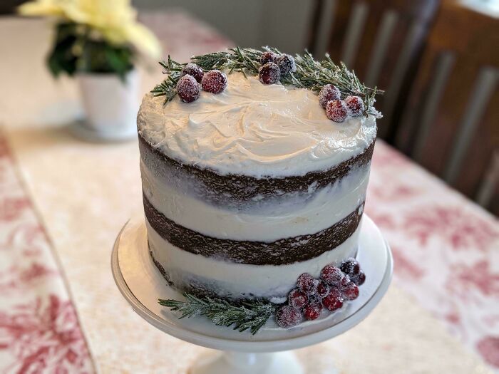 Layered cake with white frosting, topped with sugared cranberries and rosemary, showcasing bakers' culinary creativity.