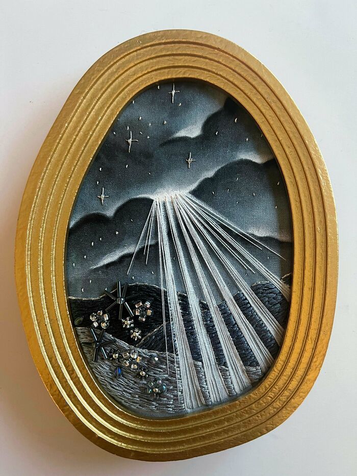 Embroidered night sky landscape with shining stars and light rays framed in a decorative gold oval frame.