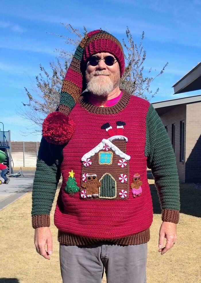 Man wearing a colorful crochet sweater and matching hat featuring a gingerbread house and Christmas decorations outdoors.