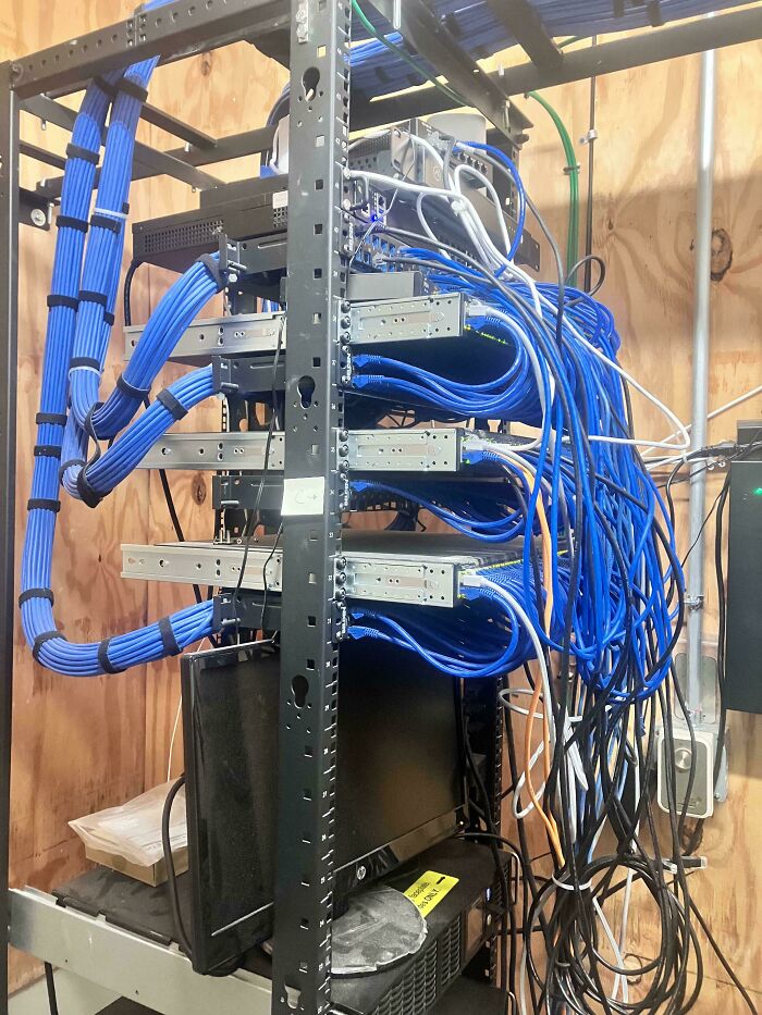 Server rack with tangled blue and gray network cables in a cluttered tech support workspace showing disorganized cable management.