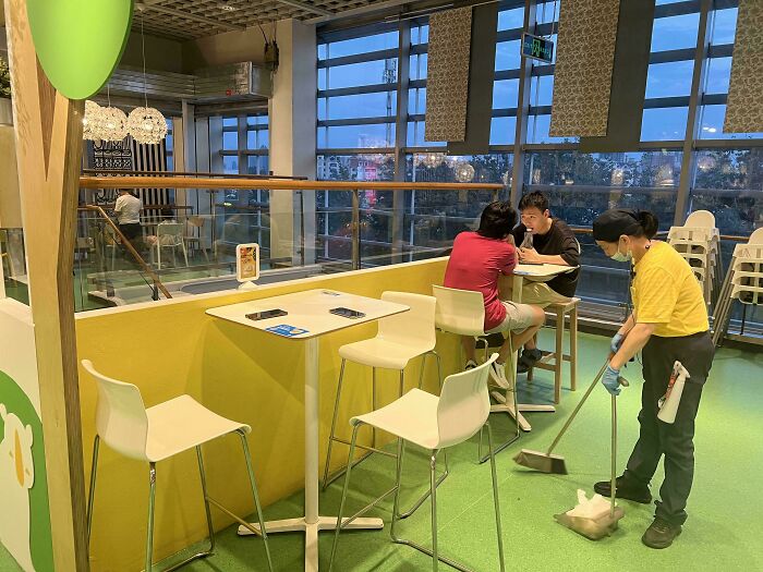 Mildly interesting scene in a café: A person sweeps the floor while two others sit at a table by large windows.