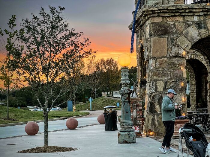 A person stands near a stone building at sunset, next to a vintage lamp, creating a mildly interesting scene.