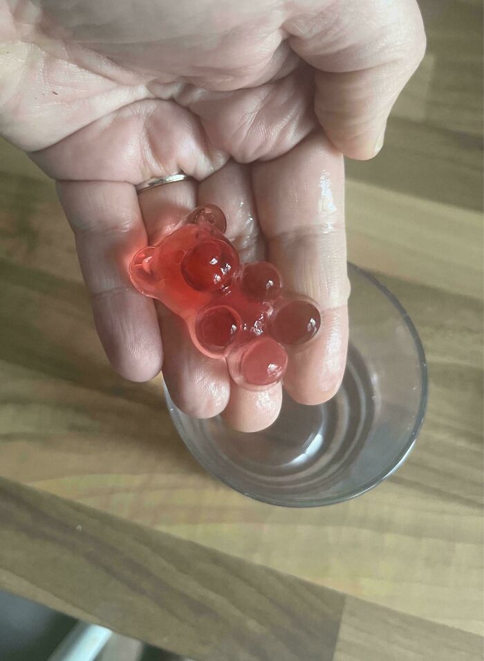 Hand holding a mildly interesting gummy bear ice cube over a glass on a wooden surface.