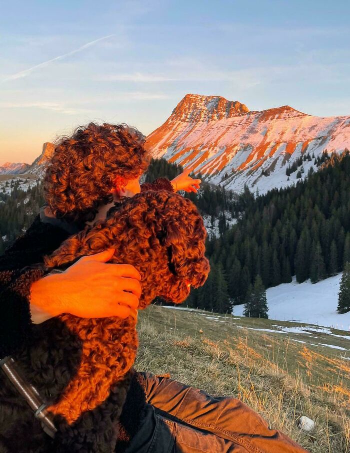 Person holding a dog while pointing at a snow-capped mountain under a sunset. Mildly interesting scene in nature.