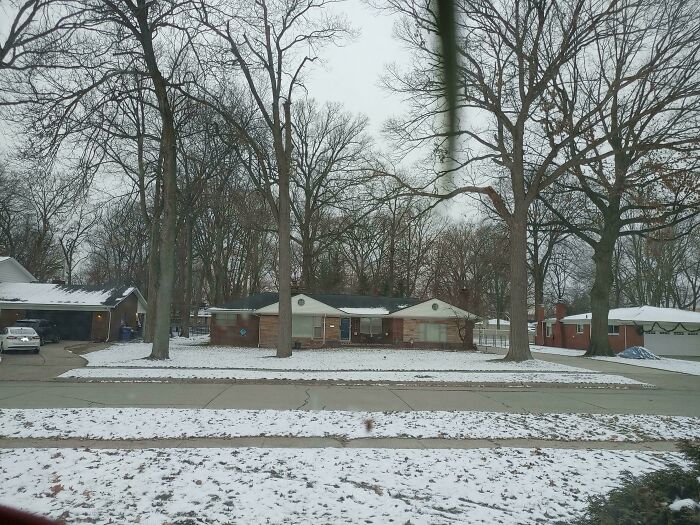 Snowy suburban street with bare trees and a single-story house, creating a mildly interesting winter scene.