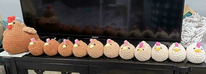 Crochet enthusiasts display a collection of handmade crocheted chickens lined up on a black cabinet beneath a TV.