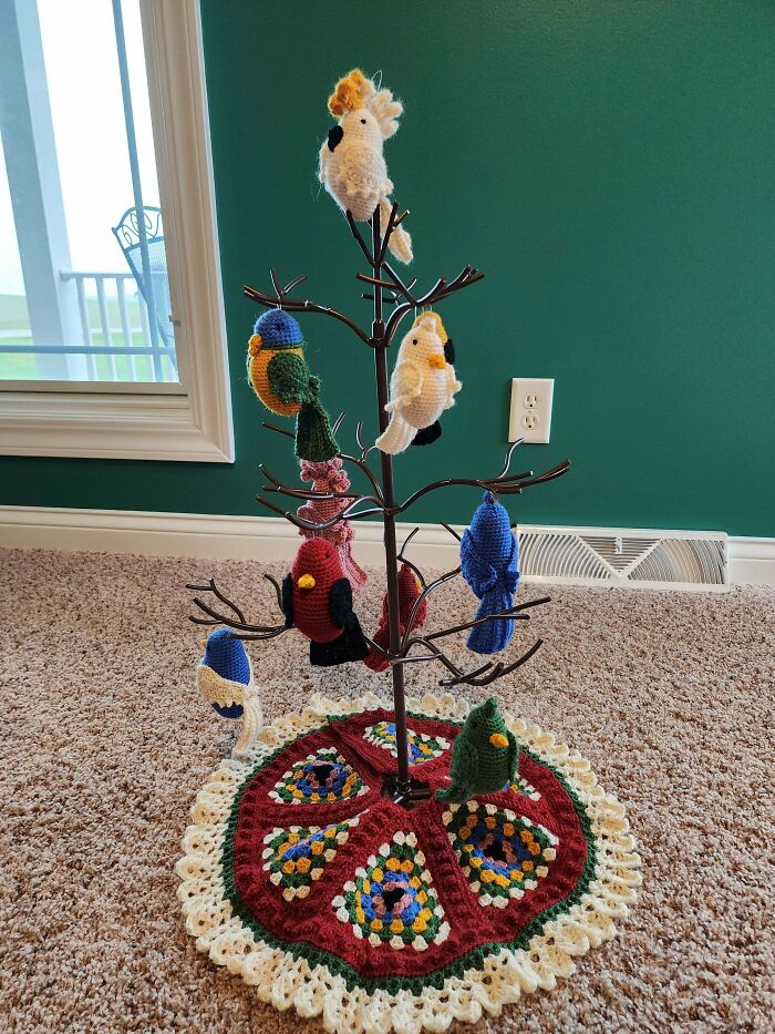 Crochet enthusiasts display colorful handmade bird decorations on a metal tree with a crocheted base in a home setting.