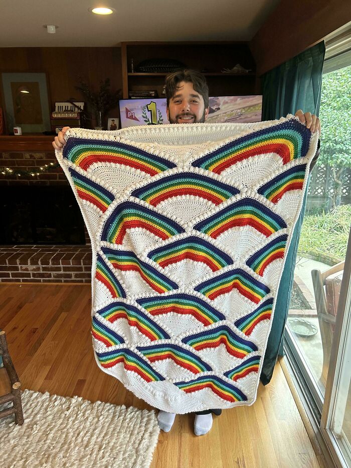 Person holding a colorful crochet blanket with rainbow patterns, showcasing a proud crochet project indoors.