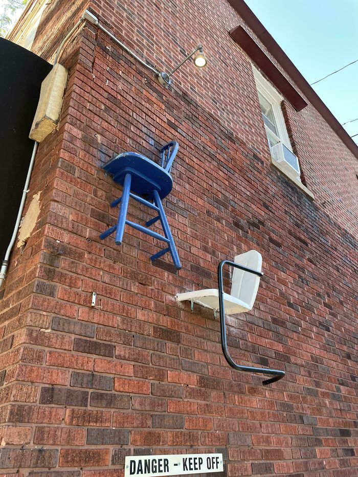 Chairs mounted on a brick wall, creating an illusion like bad renders, with a "Danger - Keep Off" sign below.