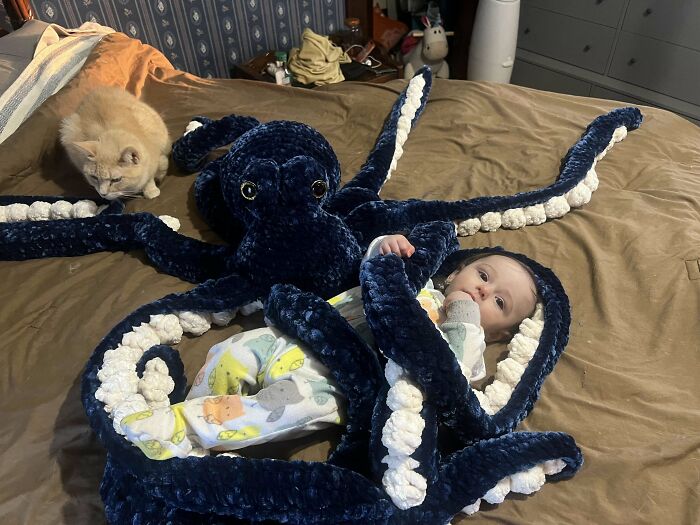 Baby wrapped in a large crochet octopus blanket with a cat nearby, showcasing proud crochet enthusiasts’ project.