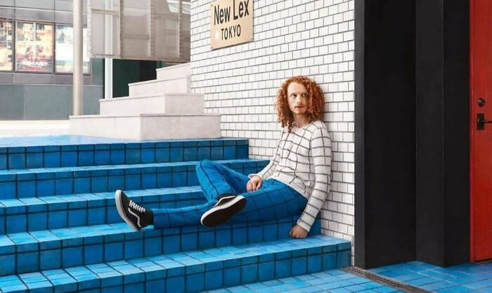 Person with curly hair in striped shirt and blue pants sitting on blue tiled steps near New Lex Tokyo sign, fascinating scene.