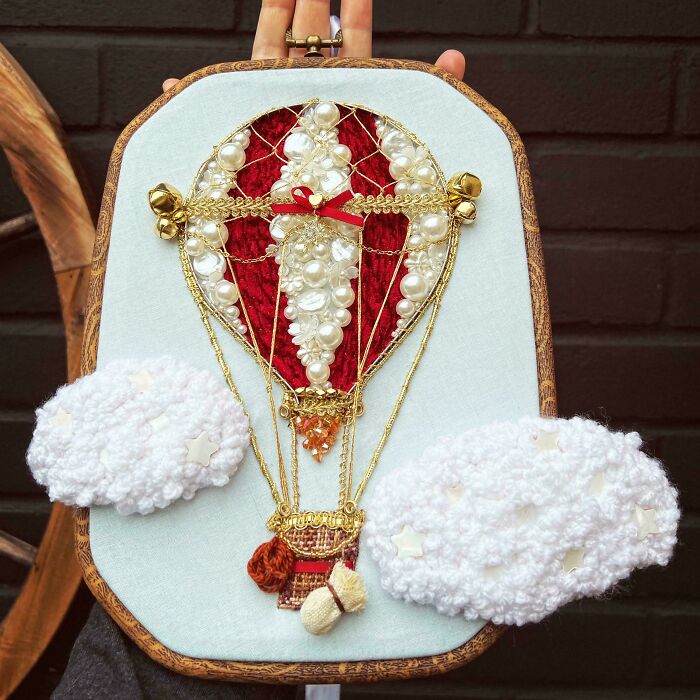 Hand holding embroidery hoop with beautiful hot air balloon design and textured clouds in embroidery designs.