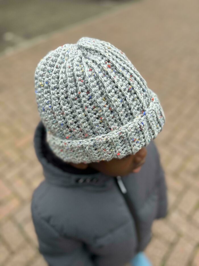 Child wearing a cozy grey crochet hat with multicolored specks, showcasing a proud crochet project with detailed stitching.