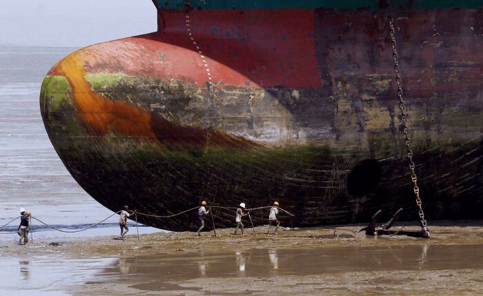 Workers pull a massive ship anchor in a muddy shore showcasing the extreme scale for megalophobia enthusiasts.