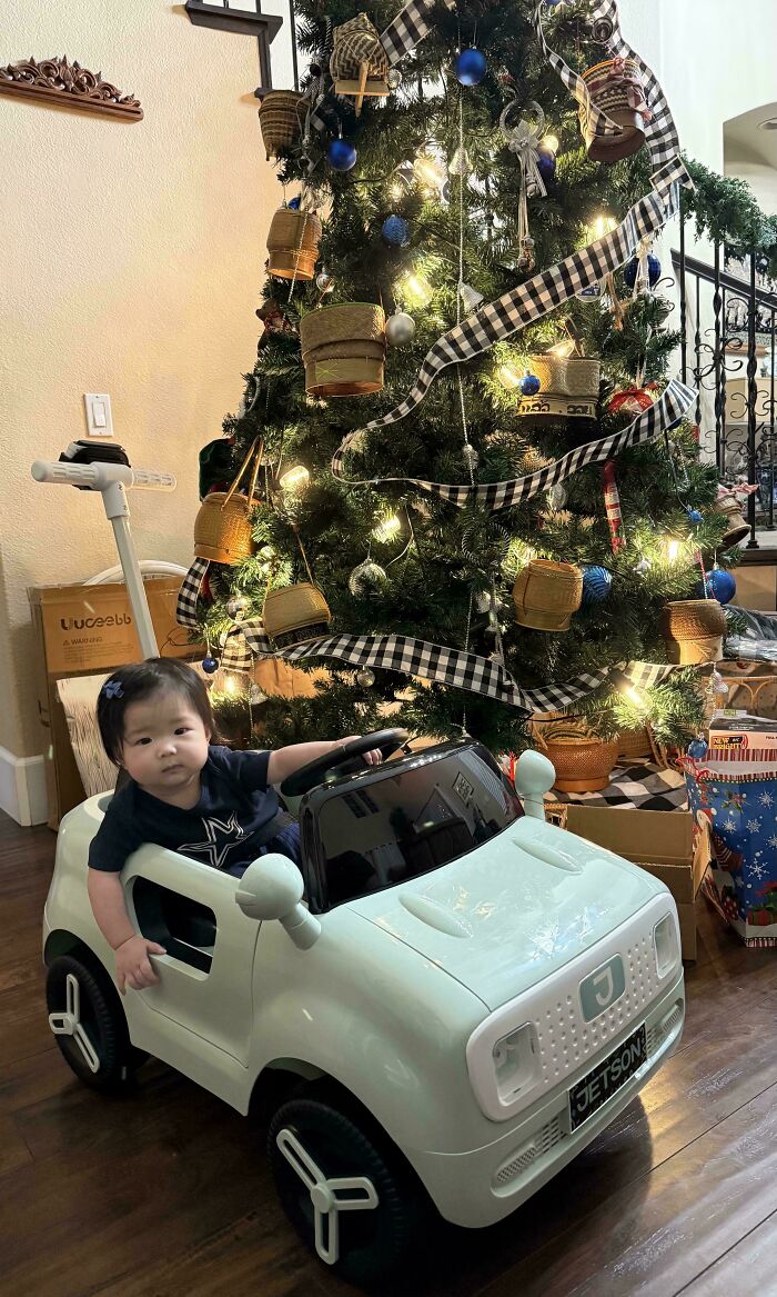 Child in white toy car by a Christmas tree, representing wholesome dad moments.