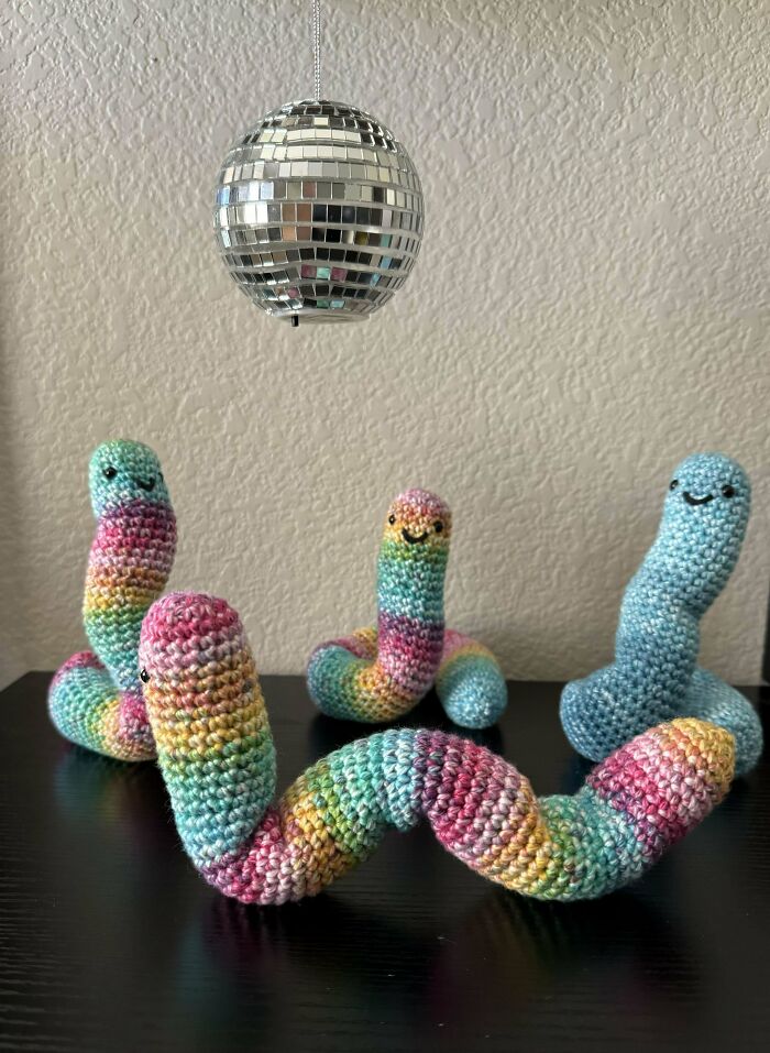 Colorful crochet worms with smiling faces displayed under a small disco ball on a dark surface, showcasing crochet projects.
