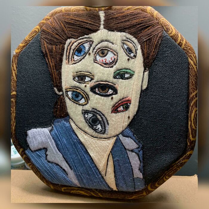 Embroidered surreal portrait featuring multiple colorful eyes on a stitched face in a decorative wooden hoop frame.