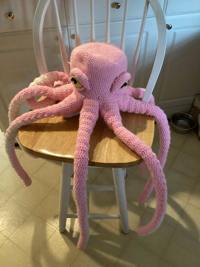 Pink crocheted octopus with detailed eyes and eight long tentacles displayed on a wooden chair by crochet enthusiasts.