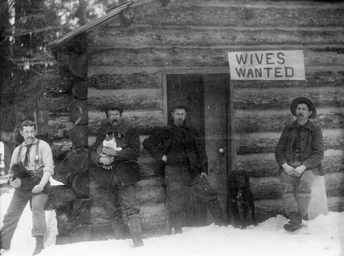 Four men outside a log cabin in snow with a sign reading "Wives Wanted" capturing fascinating historical moments.