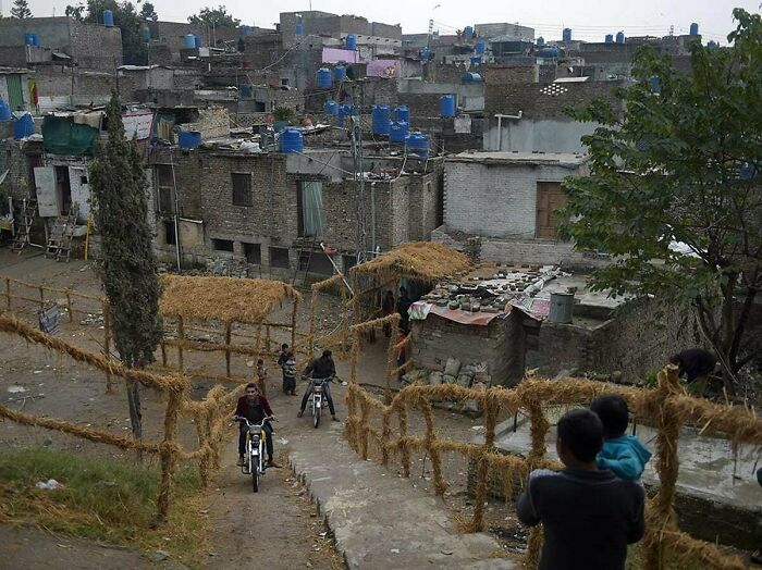 Urban hell scene with densely packed houses, dirt paths, and people on motorcycles, surrounded by makeshift barriers.