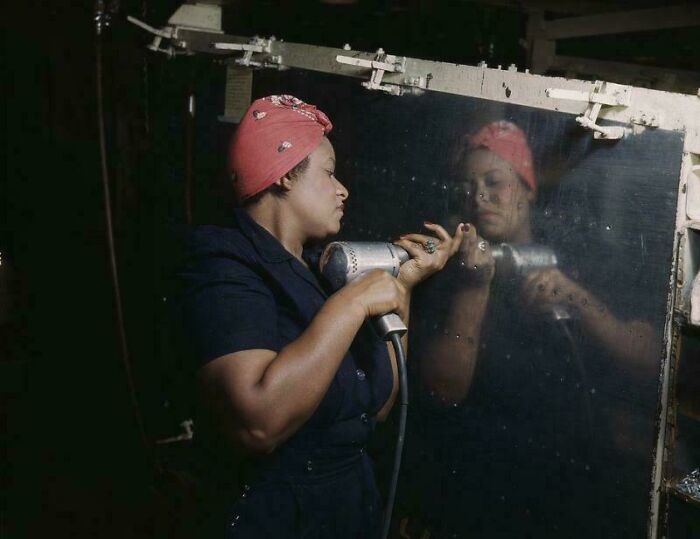 Woman in a 1940s factory setting using a drill, wearing a red headscarf, reflects the essence of historical pics.