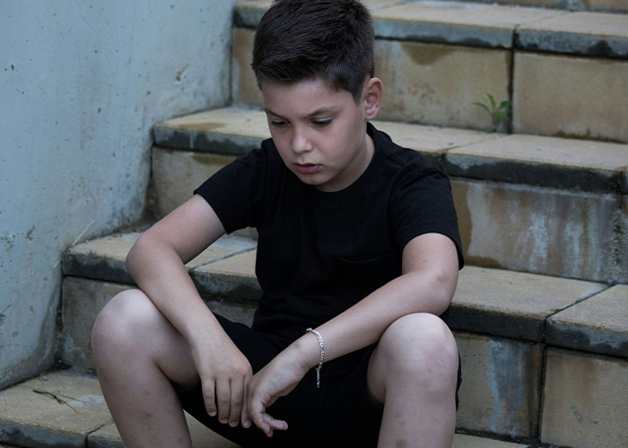 Young boy sitting on outdoor steps, looking contemplative, wearing a black outfit.