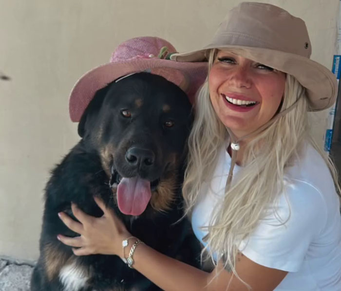 Smiling woman with long hair, wearing a hat, hugging a large dog in a pink hat, showcasing a joyful moment.