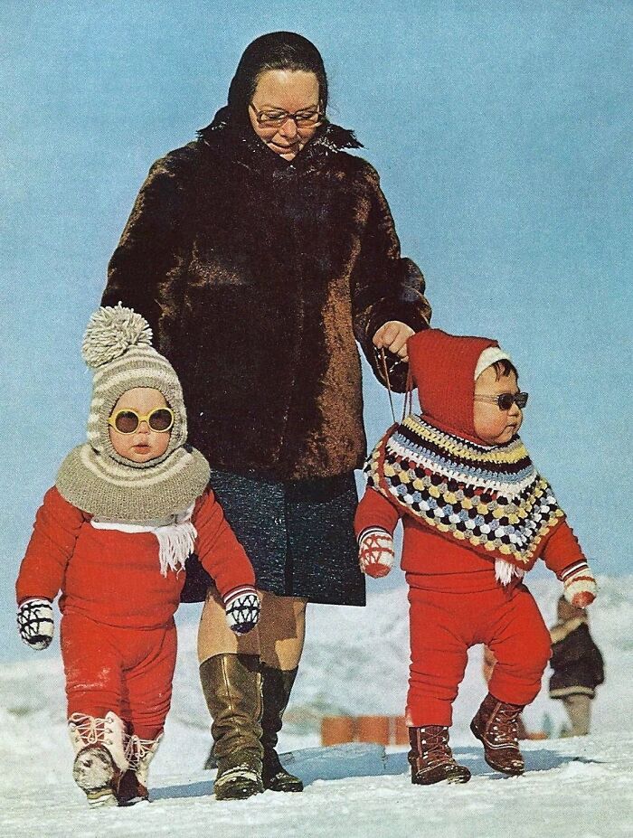 A woman with two children in winter clothing and sunglasses, walking in the snow, representing 1973 world fashion trends.