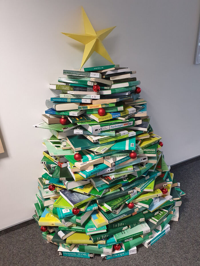 Stack of books arranged into a Christmas tree shape with a yellow star topper, decorated with red ornaments.